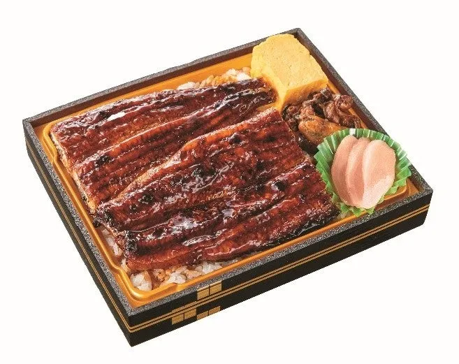 鰻の蒲焼が乗った弁当の画像です。ご飯の上に鰻が乗っており、卵焼きや漬物、その他おかずも添えられています。弁当箱は黒と金色のデザインです。