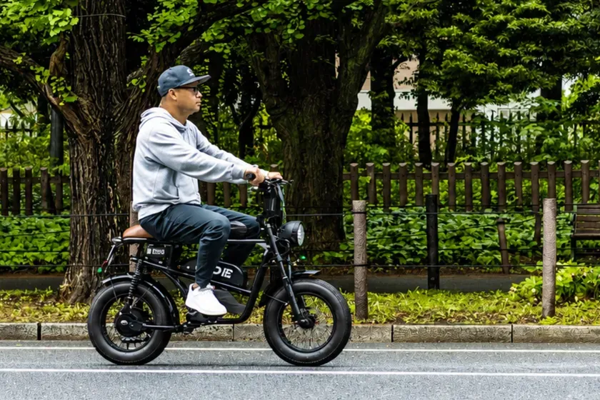 電動バイクに乗る男性の画像。男性はグレーのパーカーとキャップを着用し、サングラスをかけています。彼はバイクを運転しており、背景には木々や緑色の植物が見えます。