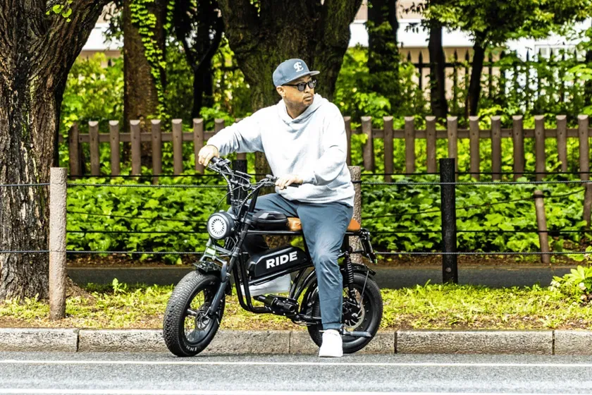 男性が電動バイクに乗っている様子を捉えた写真。彼はグレーのパーカーとキャップを着用し、サングラスをかけている。背景には緑豊かな木々とフェンスがあり、都会の風景が写し出されている。