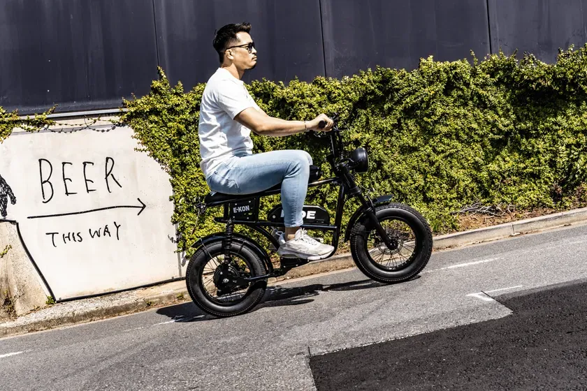 男性が電動バイクに乗っている写真。道路の脇には「BEER THIS WAY」と書かれた看板があり、緑の生け垣が背景にあります。