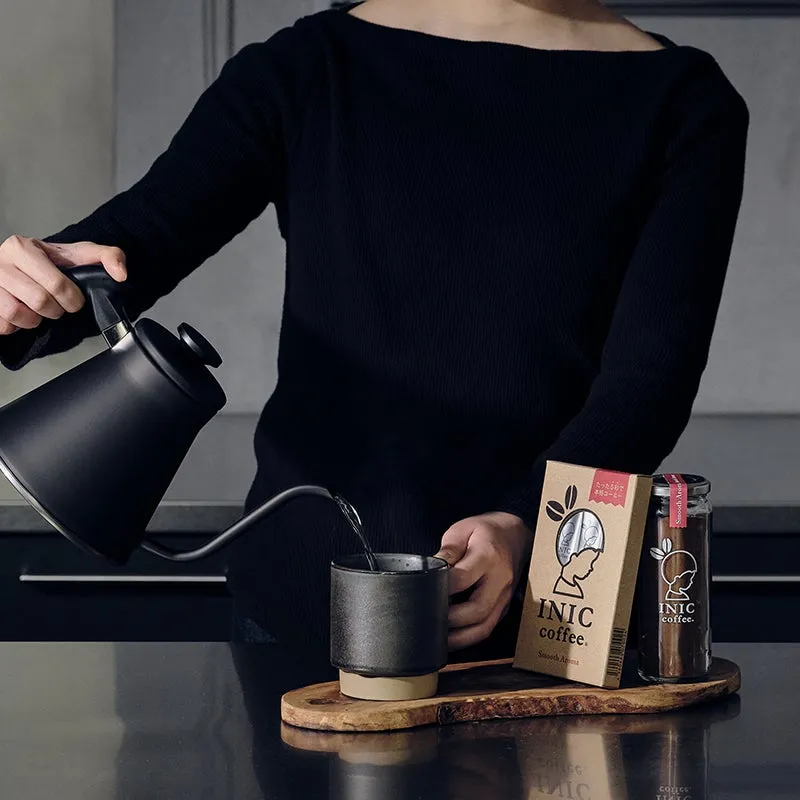 コーヒーを淹れている様子。黒い服を着た人が、黒いケトルからカップにコーヒーを注いでいる。横にはコーヒーの瓶や箱が置かれている。
