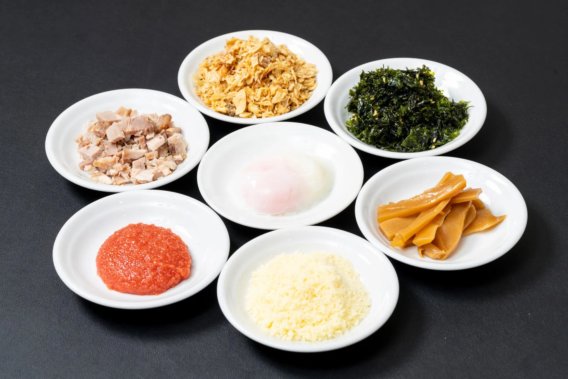 様々なラーメンのトッピングが小皿に並んだ写真。豚肉、揚げネギ、海苔、メンマ、卵、チーズ、赤いペースト状のものが見える。