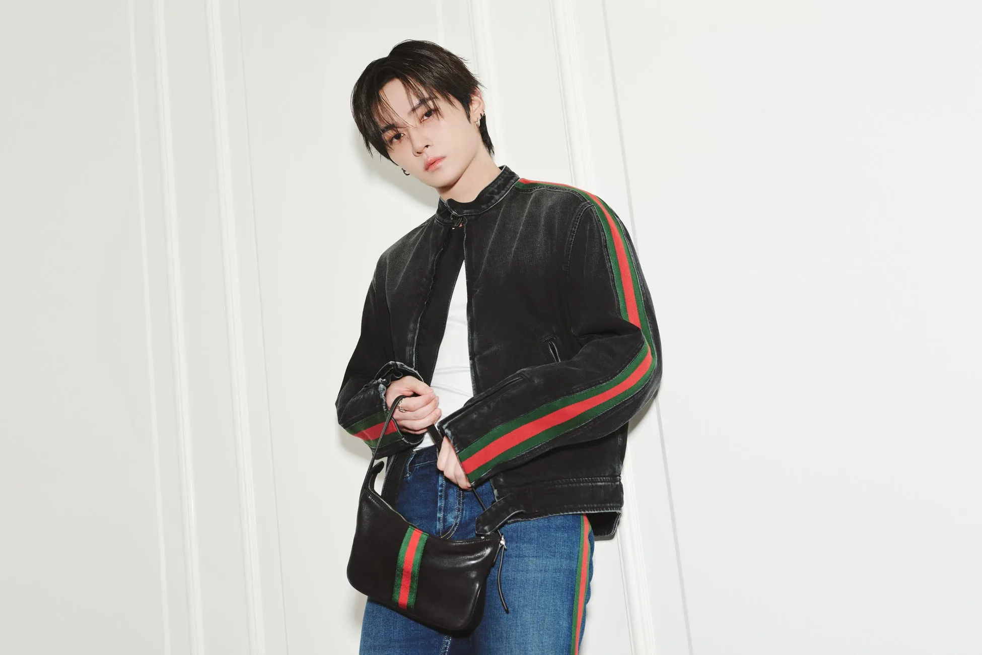 黒いジャケットとバッグを持った男性が立っている写真です。ジャケットには赤と緑のストライプが入っています。男性は白いシャツと青いジーンズを着ています。
