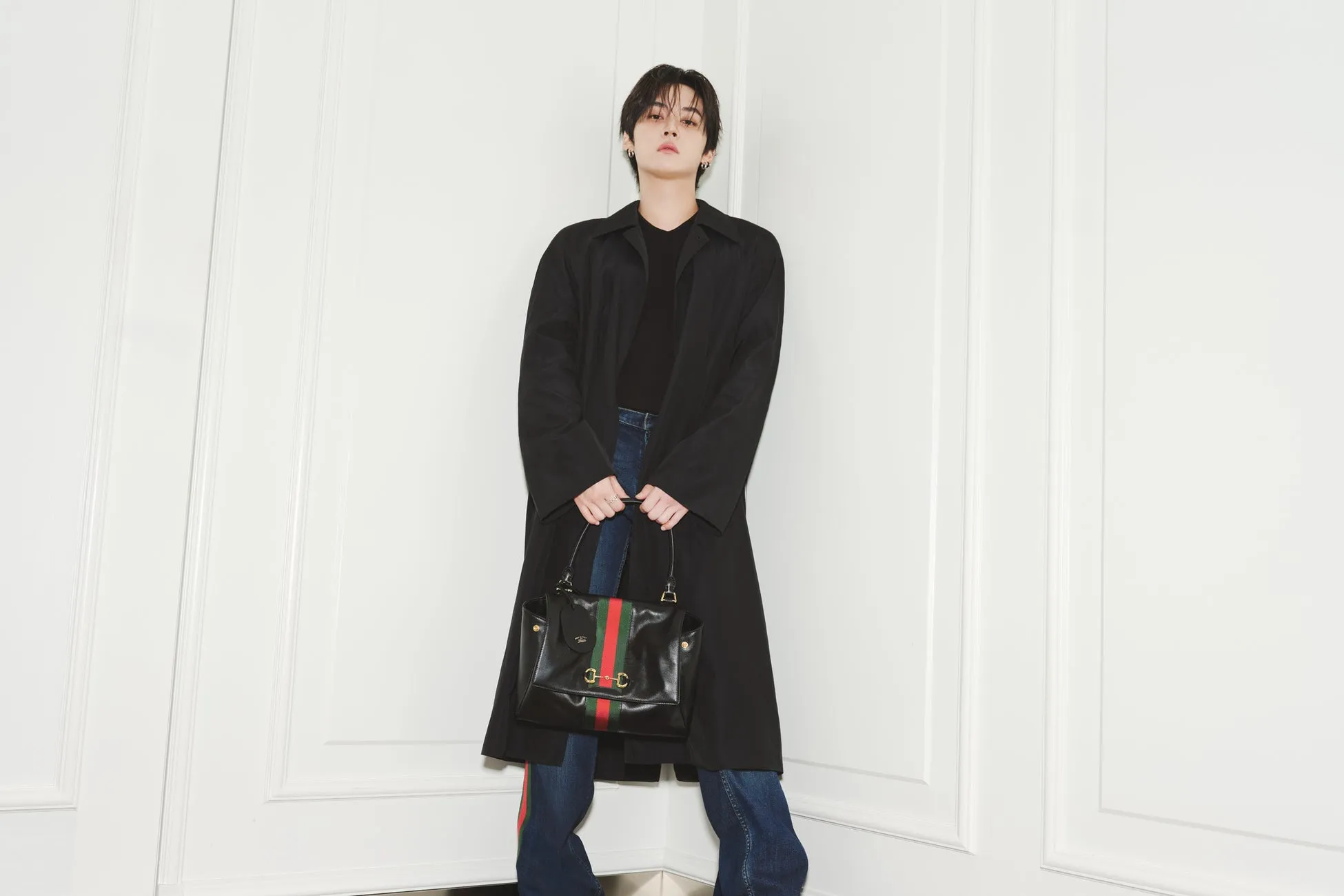 黒のコートを着た男性が黒いハンドバッグを持った写真です。背景は白い壁です。