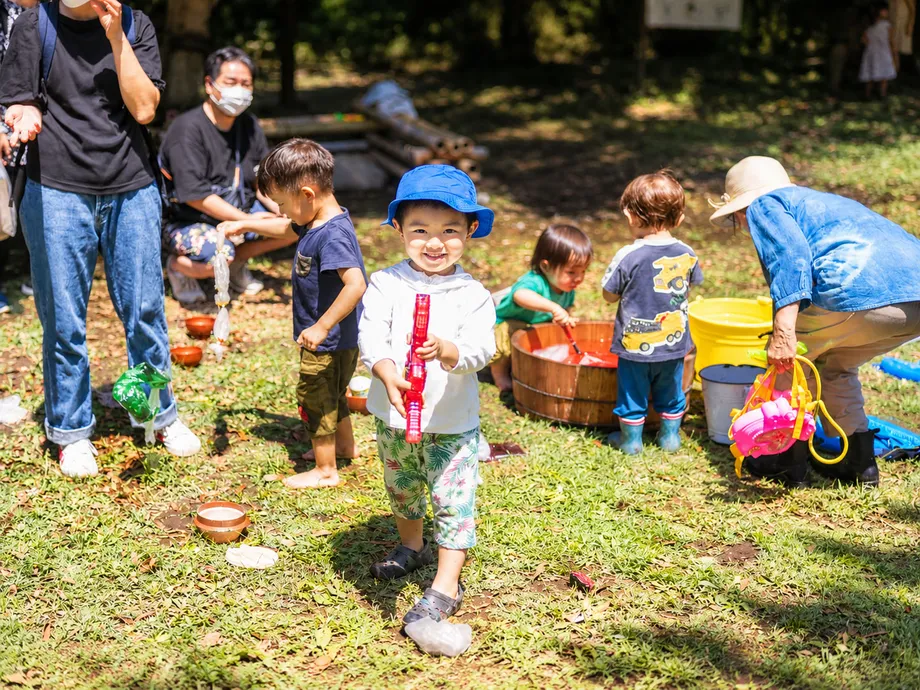 屋外で大人に見守られながら水遊びをする子どもたち