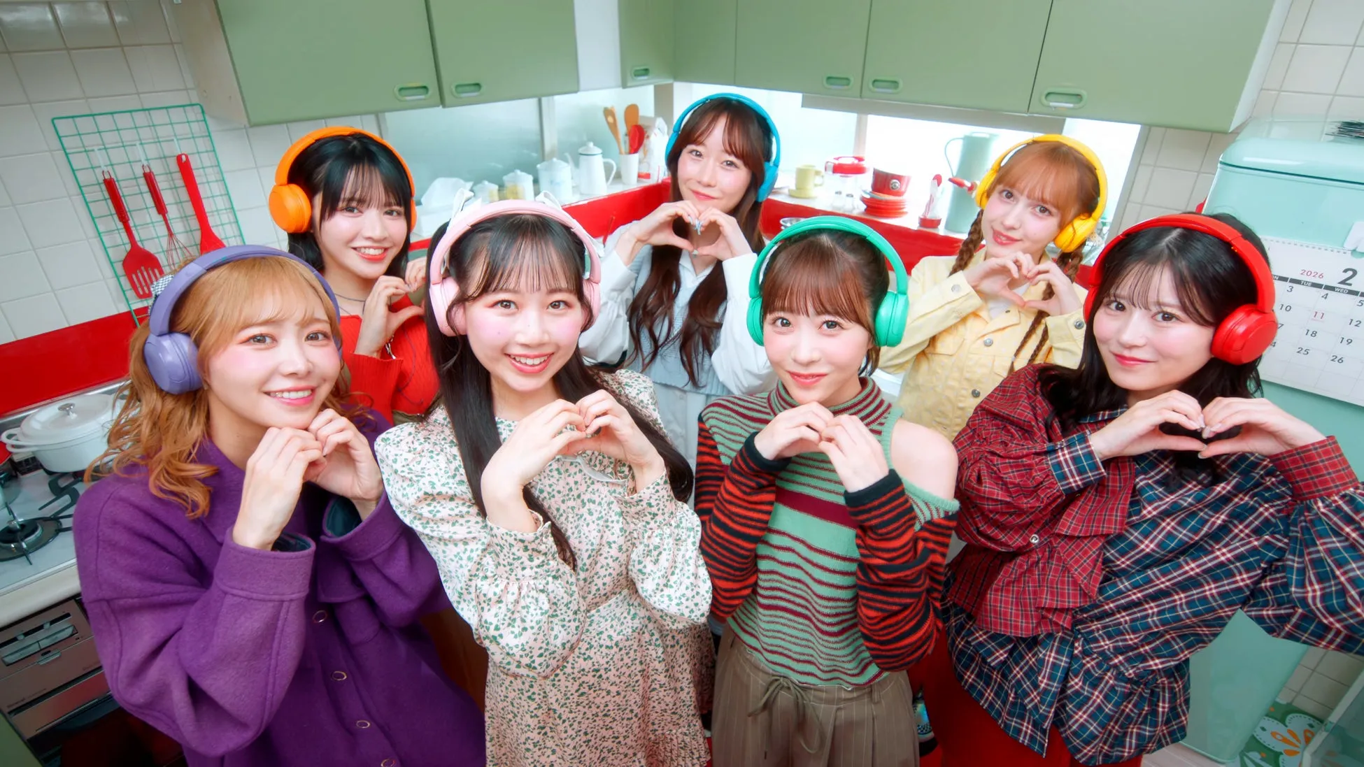 7人の女性がヘッドホンをして、ハートの形を作ってポーズをとっている写真。背景にはキッチンがあり、カレンダーも見える。