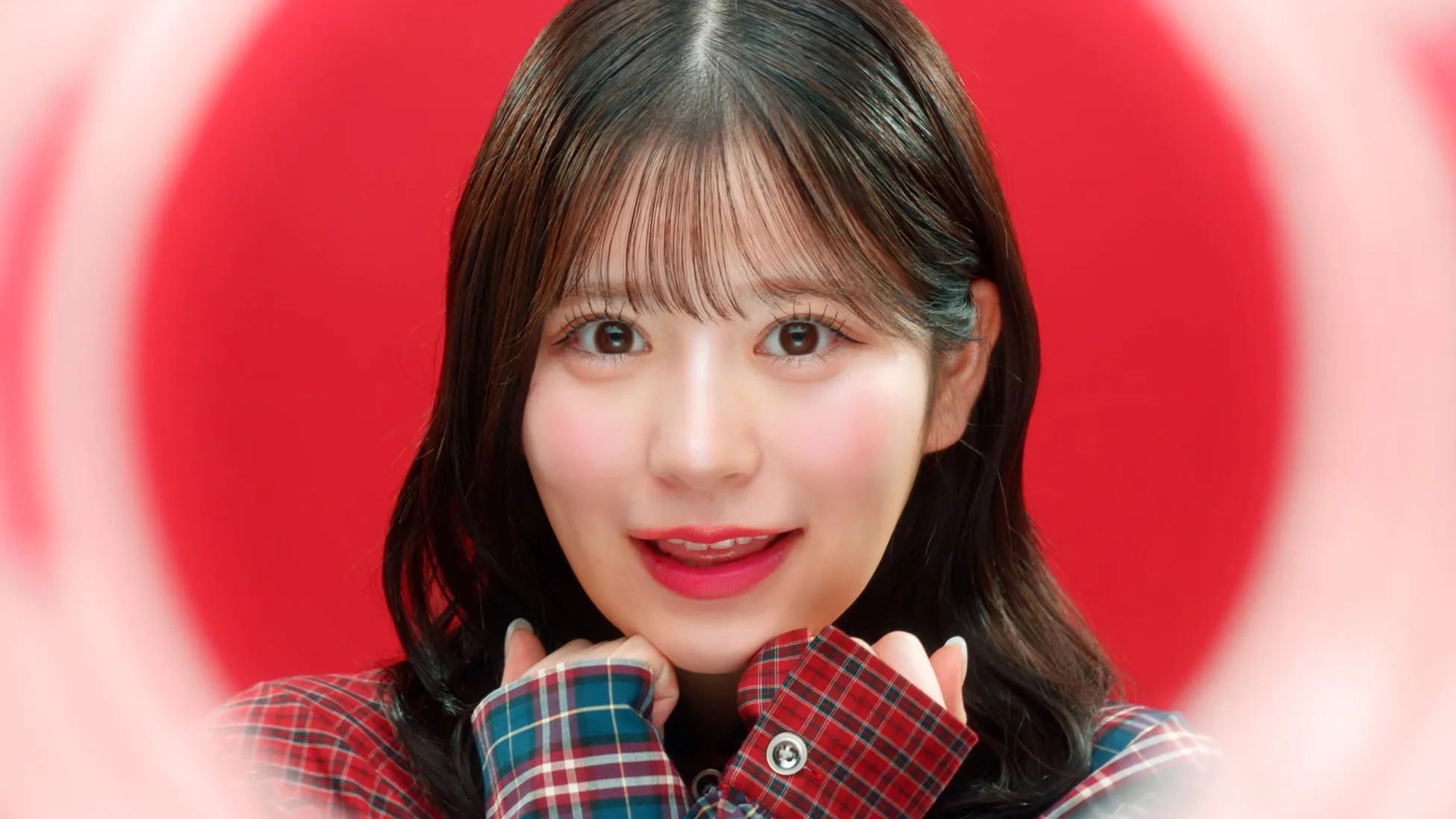赤い背景に、女性の顔がアップで写っている。女性は笑顔で、顔を両手で支えている。チェック柄のシャツを着ている。