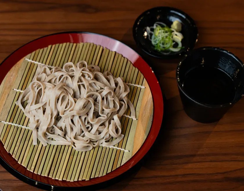 蕎麦が竹製の敷物の上に載っており、そばつゆ、ネギ、ワサビが添えられている写真。