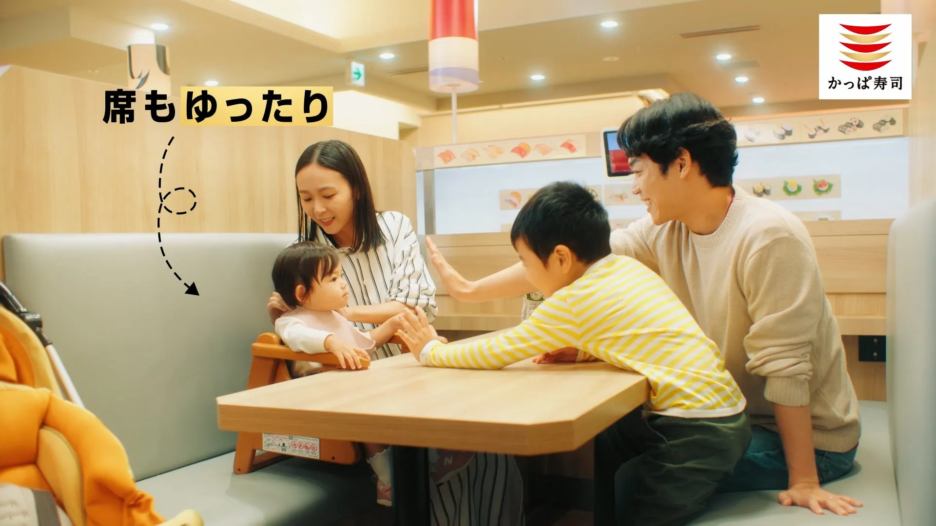 家族連れが寿司屋で食事をしている様子。子供たちはテーブルを挟んで遊んでおり、母親と父親がそれを見守っている。店のロゴが右上に見える。