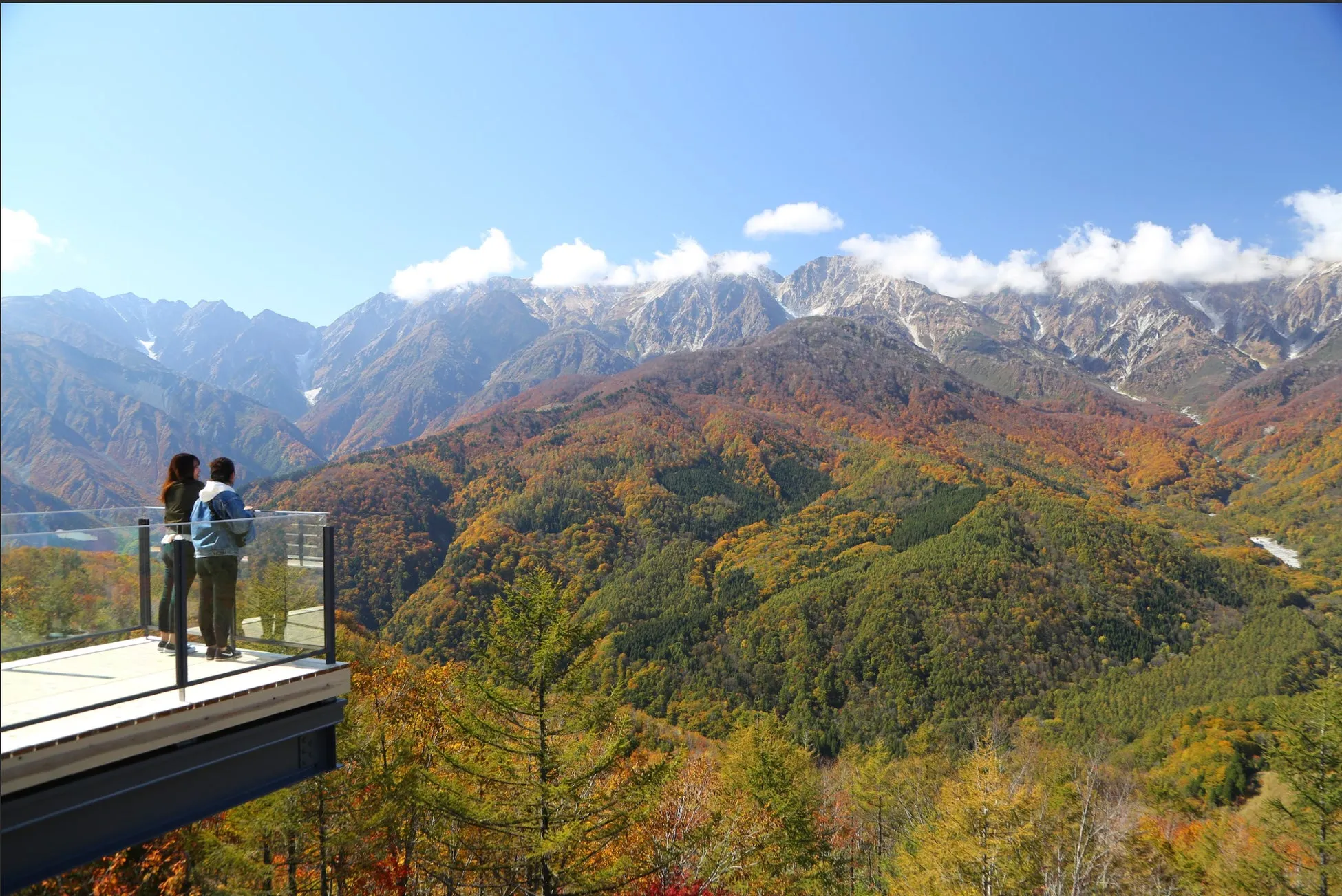 山々を見下ろす展望台に立つ2人の女性。周囲には紅葉した木々が広がり、遠くには雪を抱いた山々が連なっている。