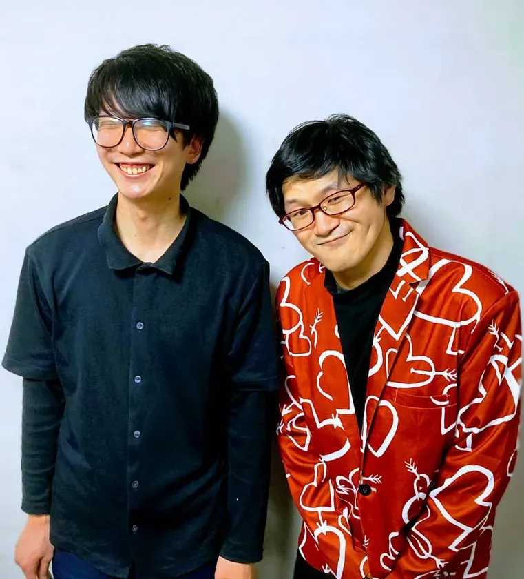 2人の男性が一緒に立って笑顔でポーズをとっている写真です。一人は黒い服を着ており、もう一人は赤いジャケットを着ています。
