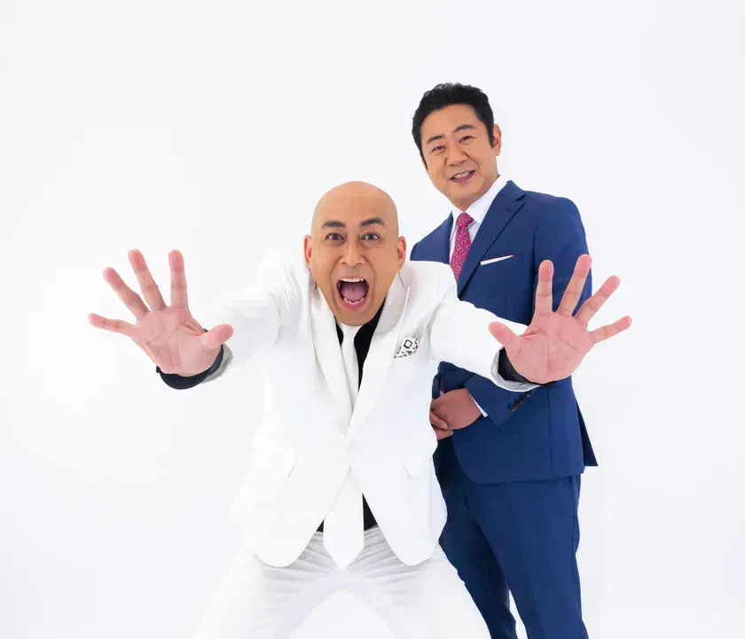 白いスーツを着た男性と、青いスーツを着た男性が写っています。白いスーツの男性は大きく口を開け、両手を前に突き出しています。青いスーツの男性はその後ろに立っています。
