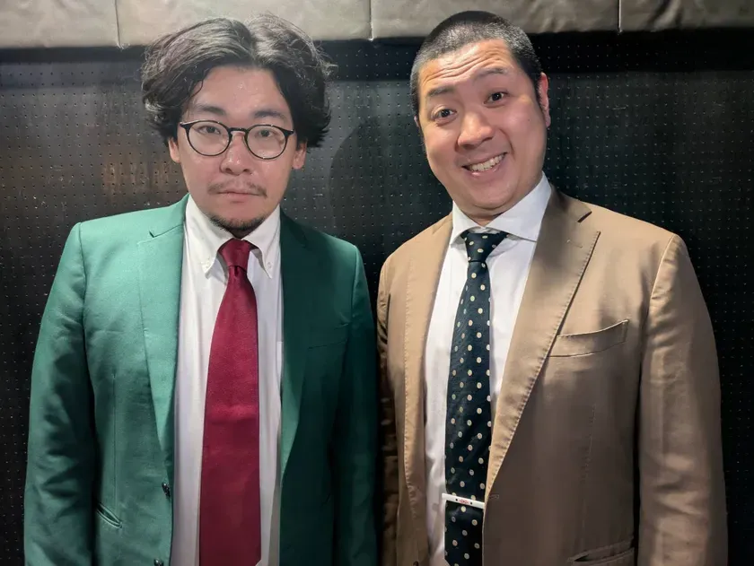 2人の男性がカメラに向かって立っている写真。左側の男性は緑色のジャケット、赤いネクタイ、眼鏡をかけています。右側の男性はベージュのジャケットと水玉模様のネクタイをしています。