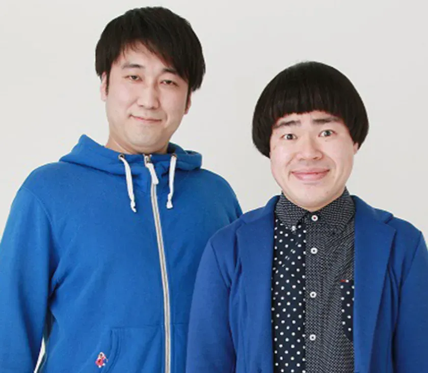 2人の男性が笑顔で立っている写真です。左側の男性は青いパーカーを着ており、右側の男性は青いジャケットを着ています。