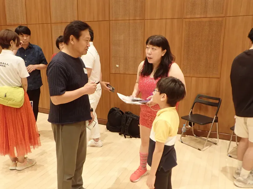 人々が会議室のような場所に集まって話している写真です。中心には、女性と男の子がおり、他の人々もいます。女性は赤いドレスを着ており、男の子は黄色いシャツを着ています。彼らは何かについて話しているように見えます。