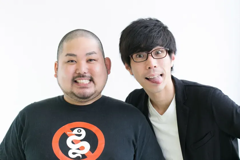 二人の男性が笑顔でポーズをとっている写真です。一人は丸刈りで、黒いTシャツを着ており、もう一人は黒髪でメガネをかけており、舌を出しています。