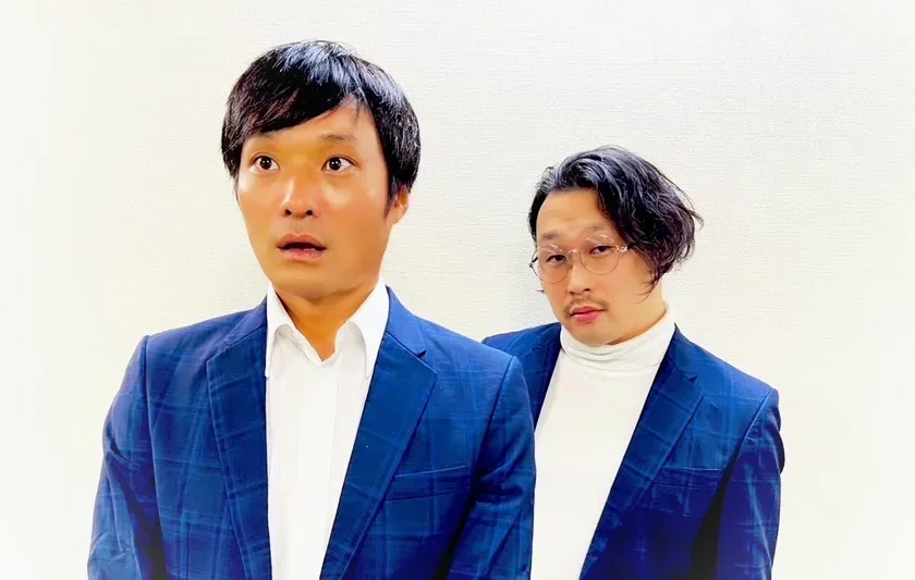2人の男性が写った写真。一人は青いチェックのスーツを着ていて、驚いたような表情をしている。もう一人は白いタートルネックと丸い眼鏡をかけている。