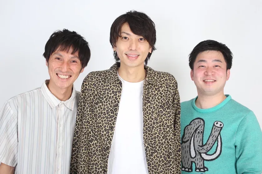 3人の男性が白い背景を前に立って笑顔でポーズをとっている写真です。真ん中の男はヒョウ柄のジャケットを着ており、左の男はストライプのシャツを着ており、右の男は象の模様のセーターを着ています。