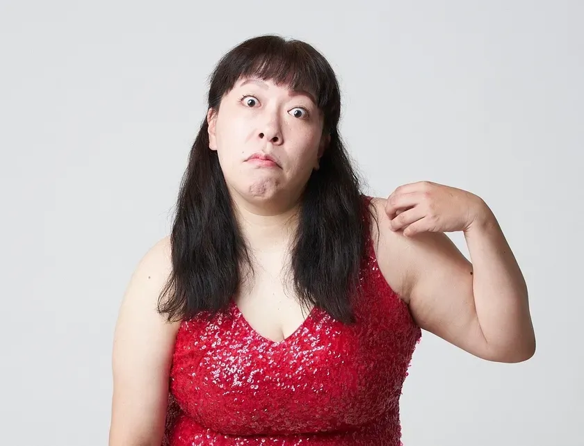 赤いスパンコール ドレスを着た女性が、驚いた表情で肩を掻いている様子を捉えた写真です。背景は白です。