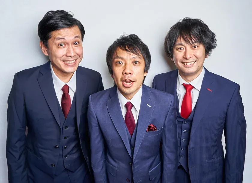 3人の男性がスーツを着て、笑顔でカメラに向かってポーズをとっている写真です。真ん中の男性は驚いたような表情で口を開けています。背景は白い壁です。