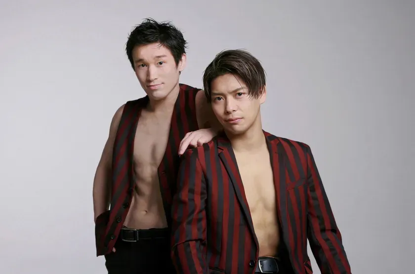 2人の男性が上半身裸で立っている写真。2人ともストライプのジャケットまたはベストを着ている。背景は白。
