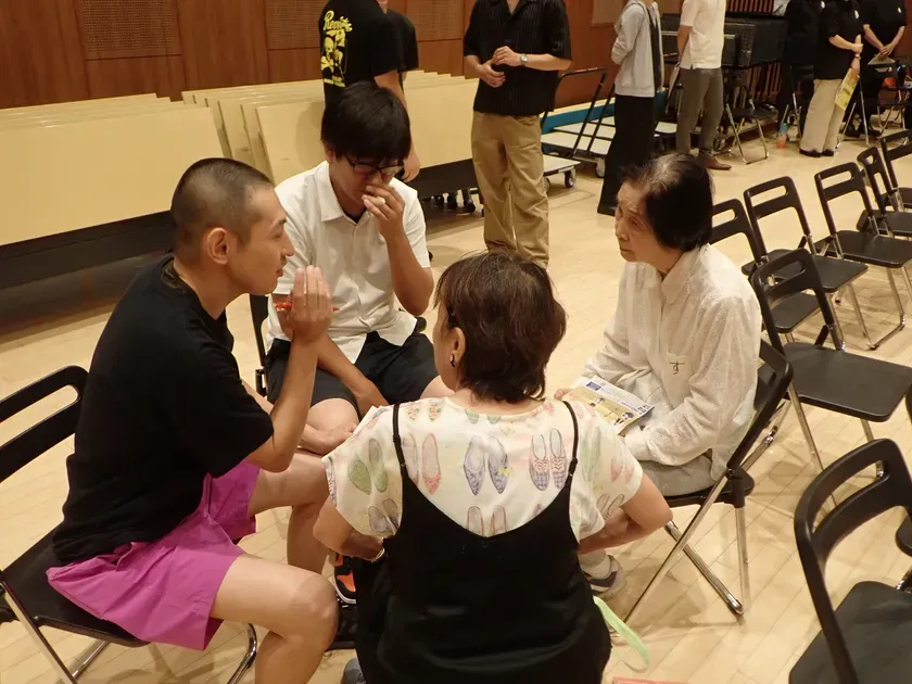 4人の人々が椅子に座って話し合っている様子。他の人々も部屋に立っている。