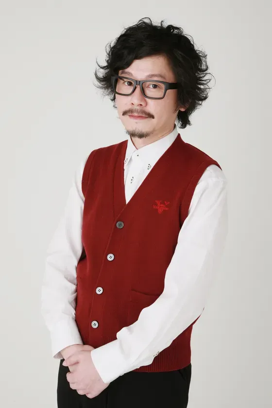 メガネをかけた男性が、赤いベストと白いシャツを着て、腕を組んで立っている様子を写した写真です。背景は白です。