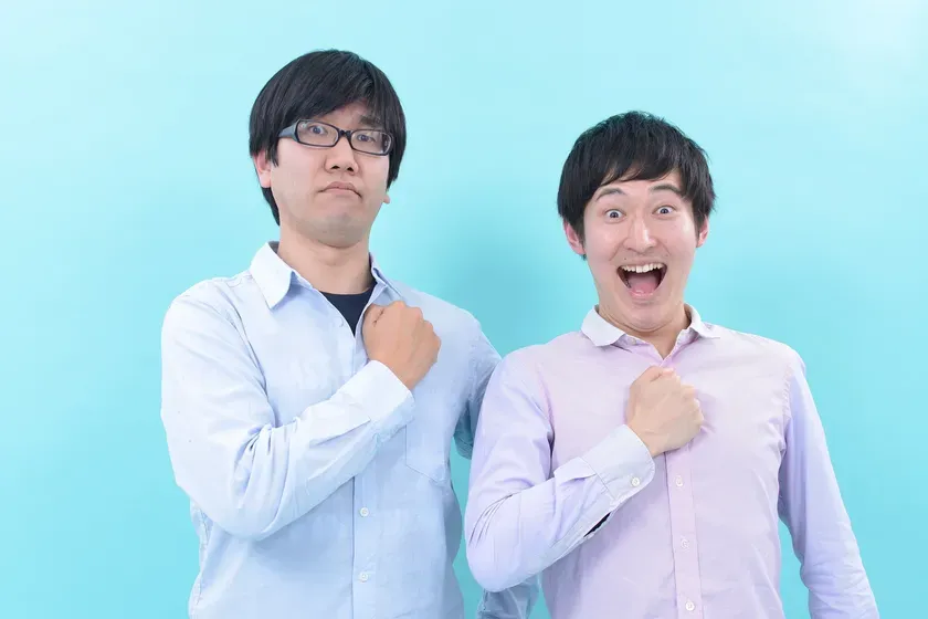 2人の男性が胸に手を当ててポーズをとっている写真。一人は眼鏡をかけ、もう一人は口を開けている。
