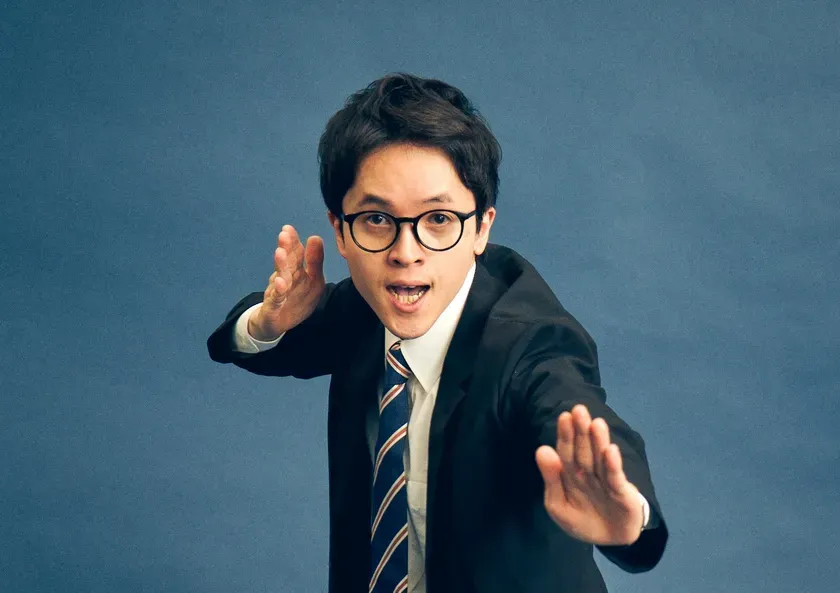 スーツ姿の男性が、両手を広げて何かを説明しているような様子を写した写真です。彼はメガネをかけており、ネクタイを締めています。背景は青色です。