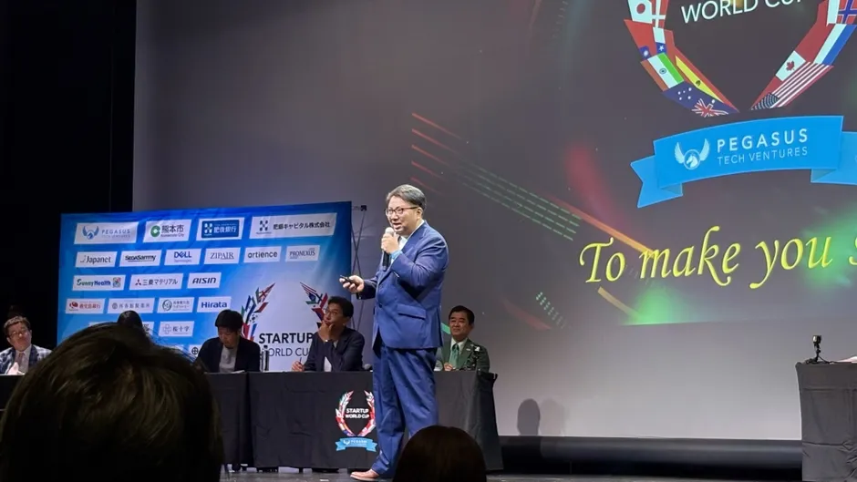 男性がステージでマイクを持って話している。背景には、企業のロゴや「STARTUP WORLD CUP」の文字がデザインされたスクリーンがある。