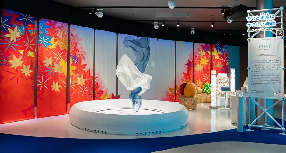 展示会場の様子。白い円形の台の上に、青と白の布が舞っているように展示されている。背景には紅葉の絵が描かれたパネルが並び、様々な展示物も見える。