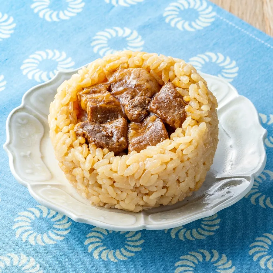 ご飯で肉を包んだ料理の画像です。白い皿の上に、ご飯で包まれた肉が乗っています。背景には青い布が敷かれています。