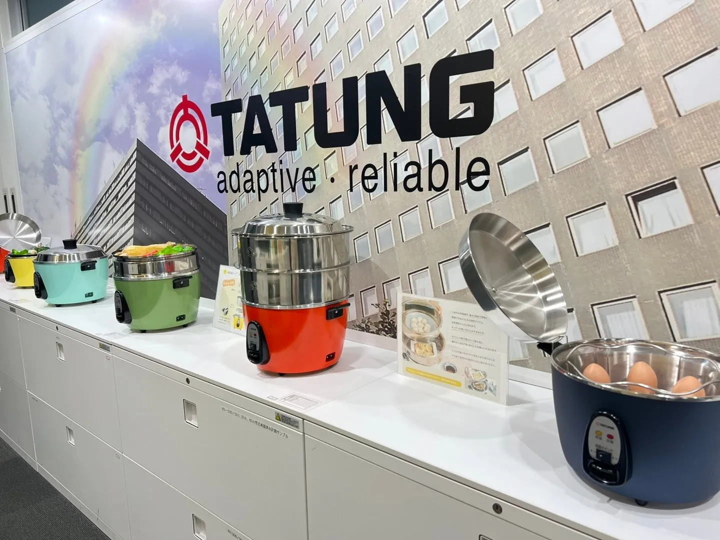 様々な色の電気蒸し器が並んだ展示会の写真。背景にはTATUNGのロゴとスローガンが描かれています。