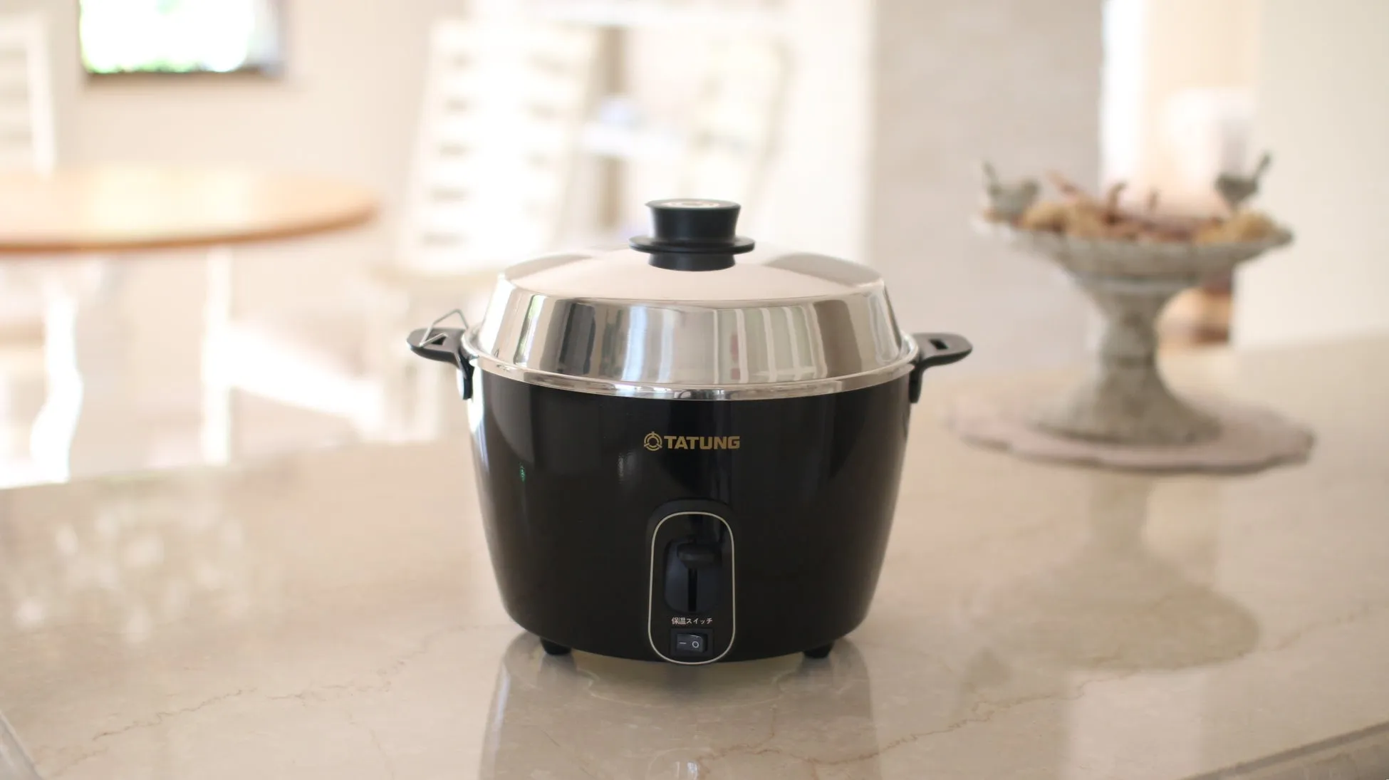 黒い炊飯器が、明るいキッチンカウンターに置かれています。炊飯器には「TATUNG」のロゴがあり、保温スイッチがあります。背景には、ぼやけたテーブルと装飾的な台座があります。