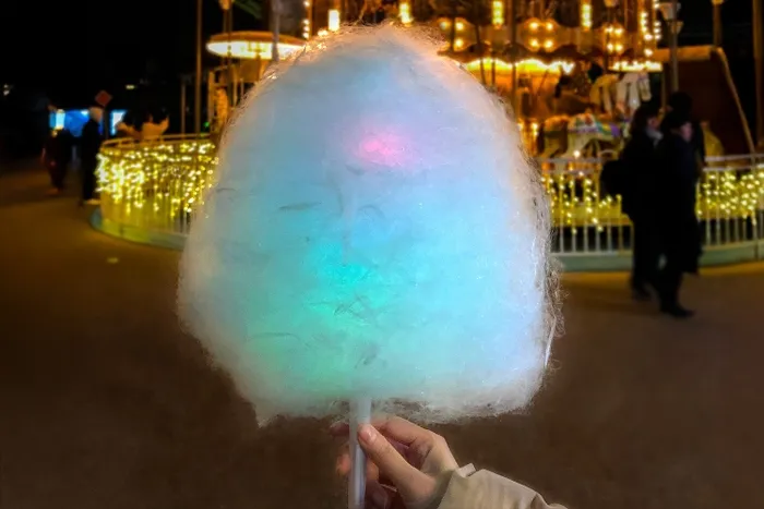 夜の遊園地で、カラフルに光る綿菓子を持った手のクローズアップ。背景には、メリーゴーランドと人影が見える。