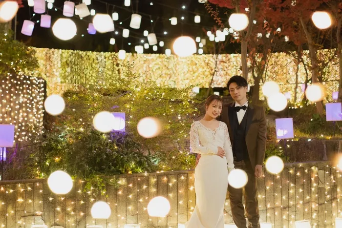 夜のライトアップされた場所で、新郎新婦が寄り添って立っている様子を捉えた写真です。背景には、丸いライトや四角いライトが多数飾られており、ロマンチックな雰囲気を醸し出しています。