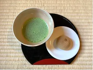 抹茶と和菓子が写った写真です。抹茶は器に注がれ、和菓子は白い小皿に載っています。背景には畳が見えます。