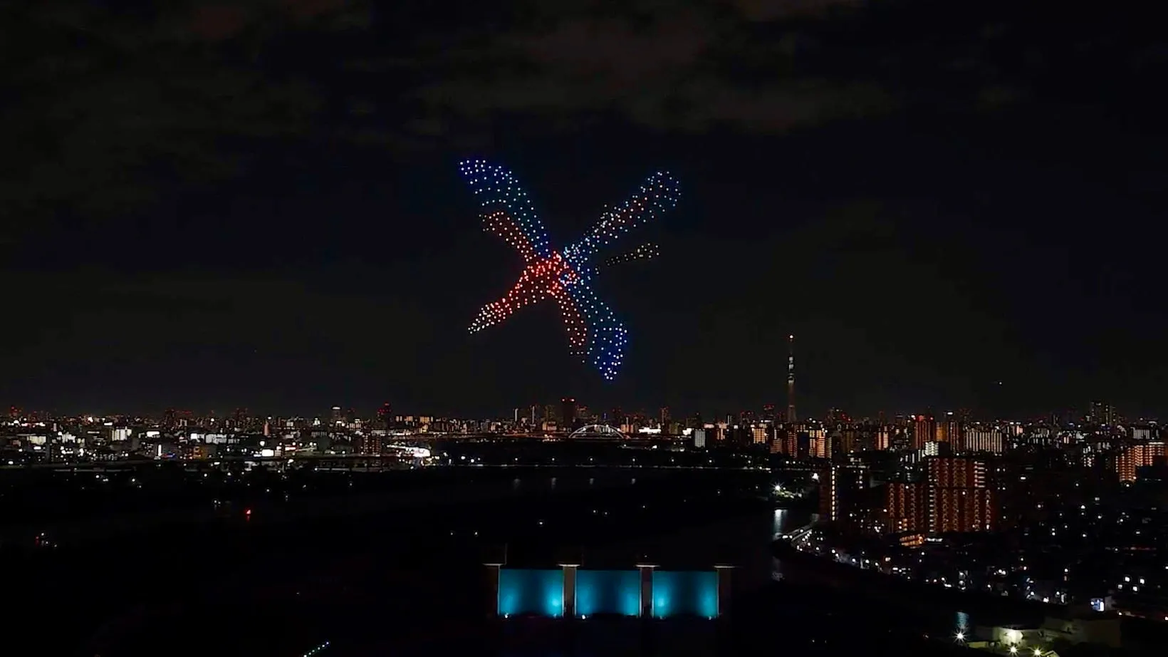 夜空に浮かぶドローンショー。ドローンが様々な色で形を作り、都市の夜景を彩っている。