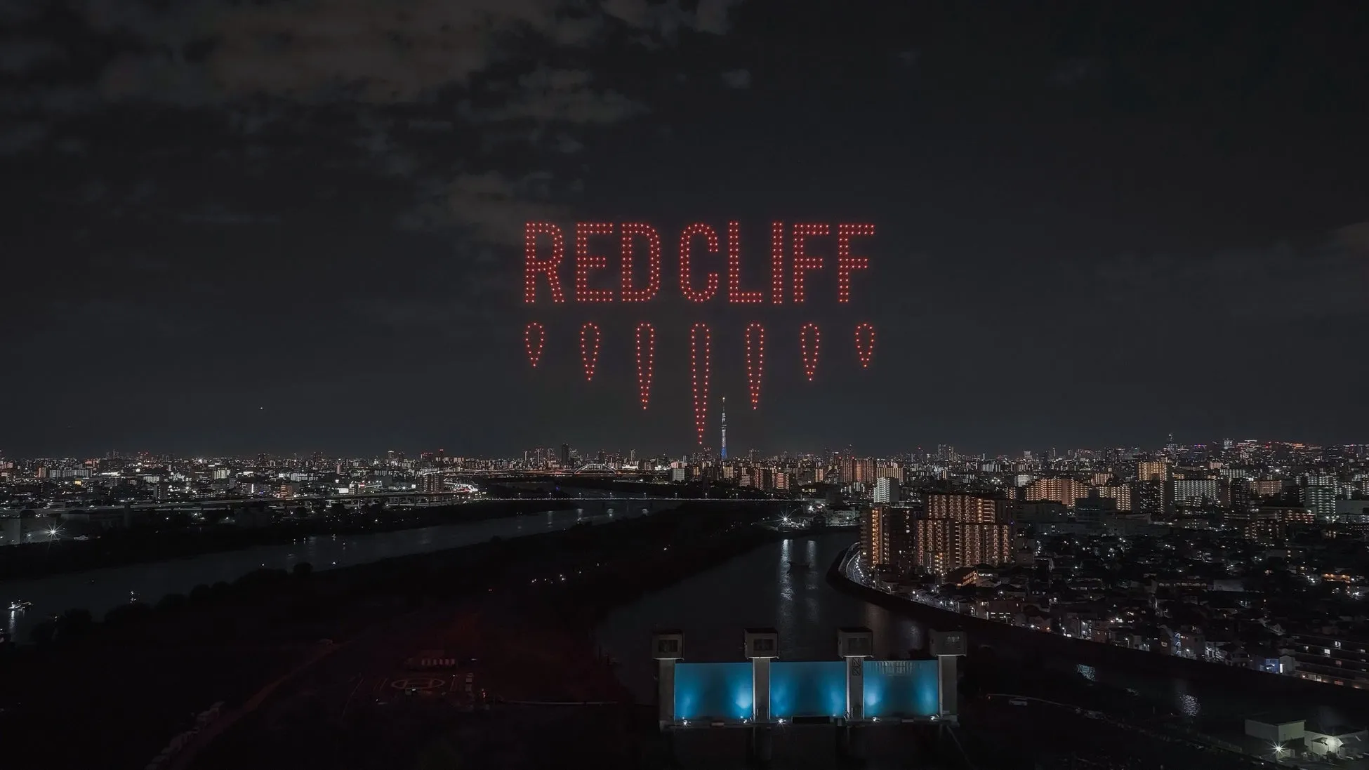 夜空に「RED CLIFF」の文字がドローンで描かれた写真。街明かりと川が写り、印象的な夜景となっている。
