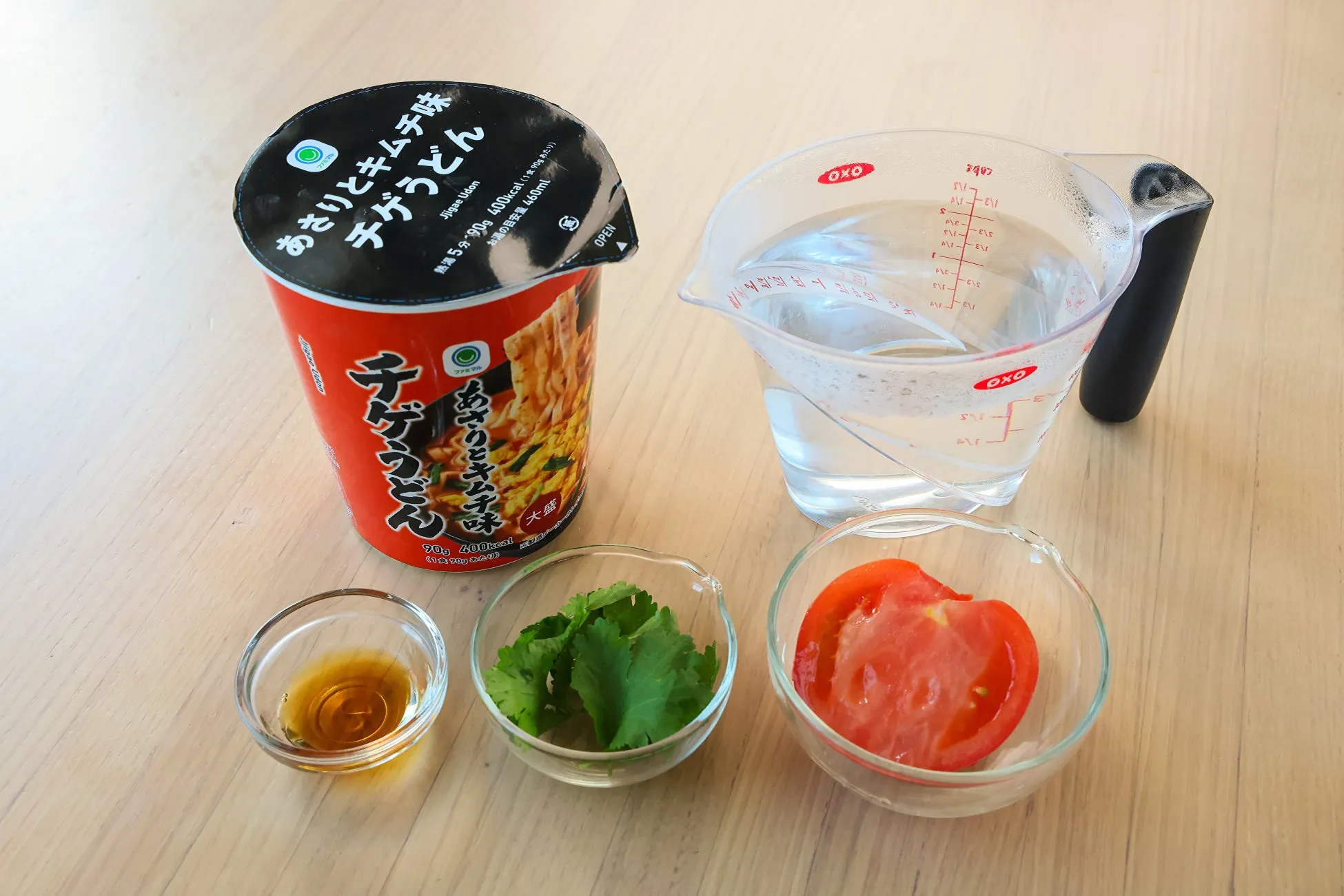 カップ麺「あさりとキムチ味 チゲうどん」と、薬味、水、計量カップ、トマトが写った写真です。