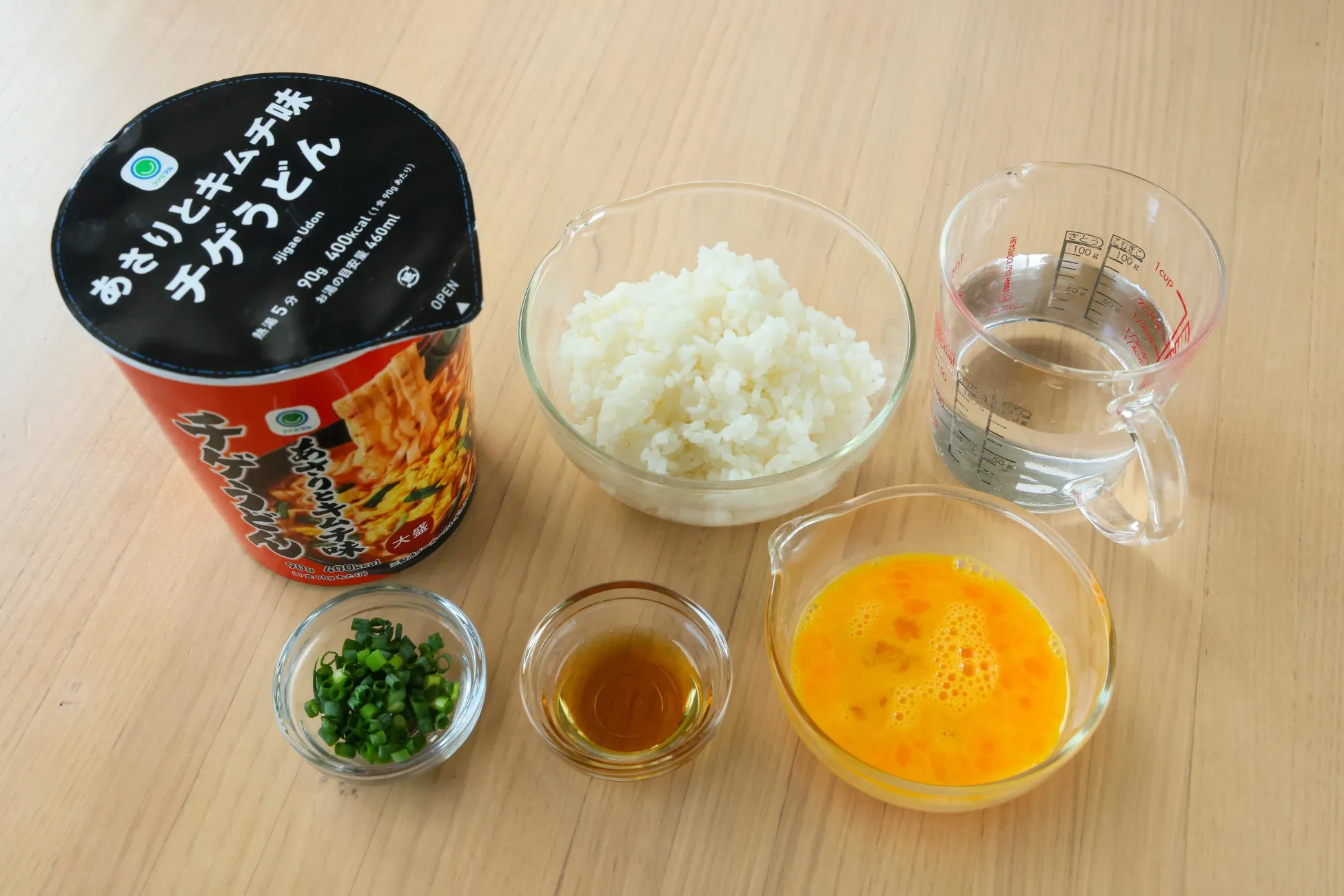 インスタントラーメンのチゲうどん、ご飯、ネギ、卵、調味料、水が入った計量カップが写っている写真。