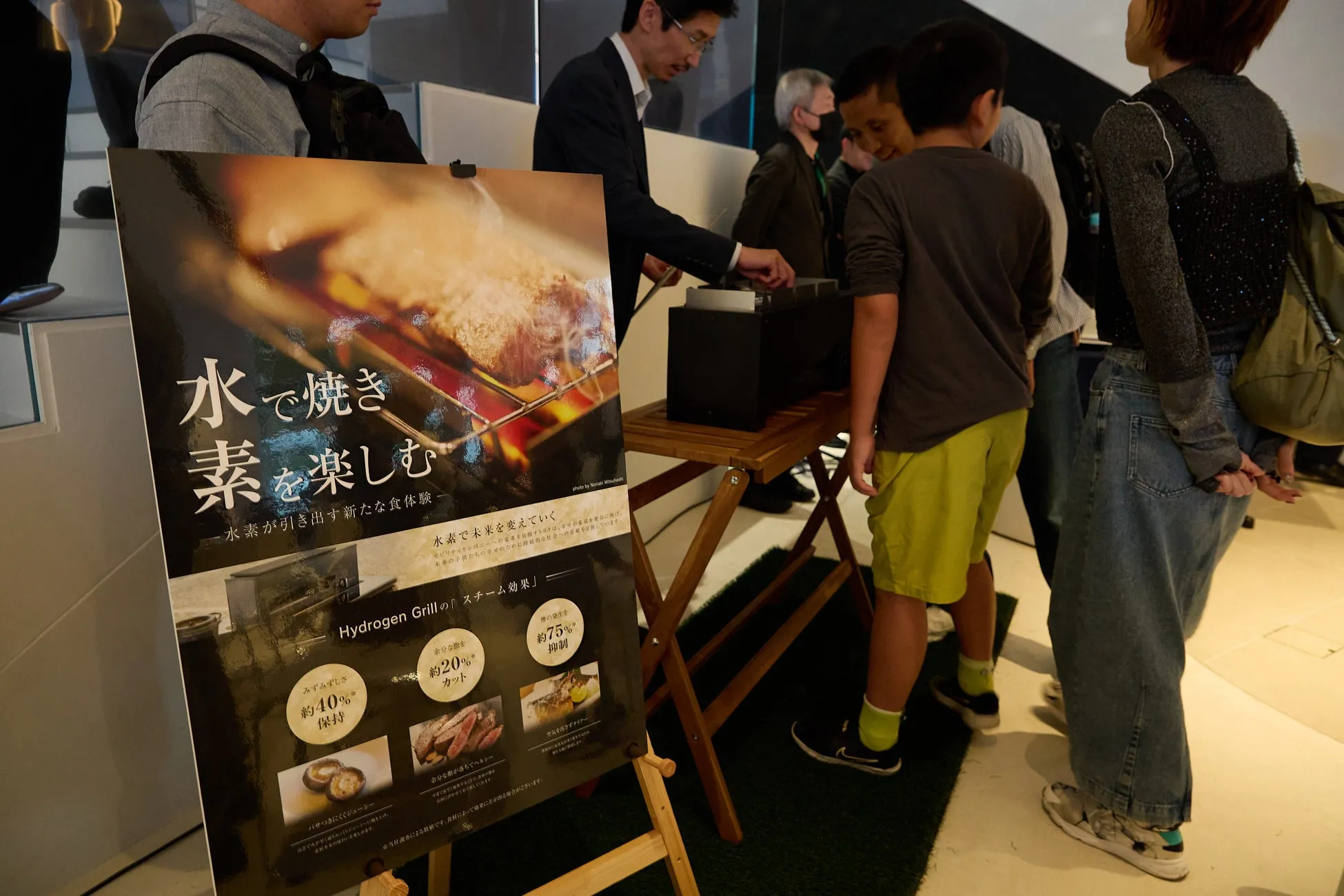 水素グリルに関する展示会で、人々が製品を試している様子が写っています。展示会には、水素グリルに関するポスターがあり、製品の利点や水素が未来を変えるというメッセージが書かれています。