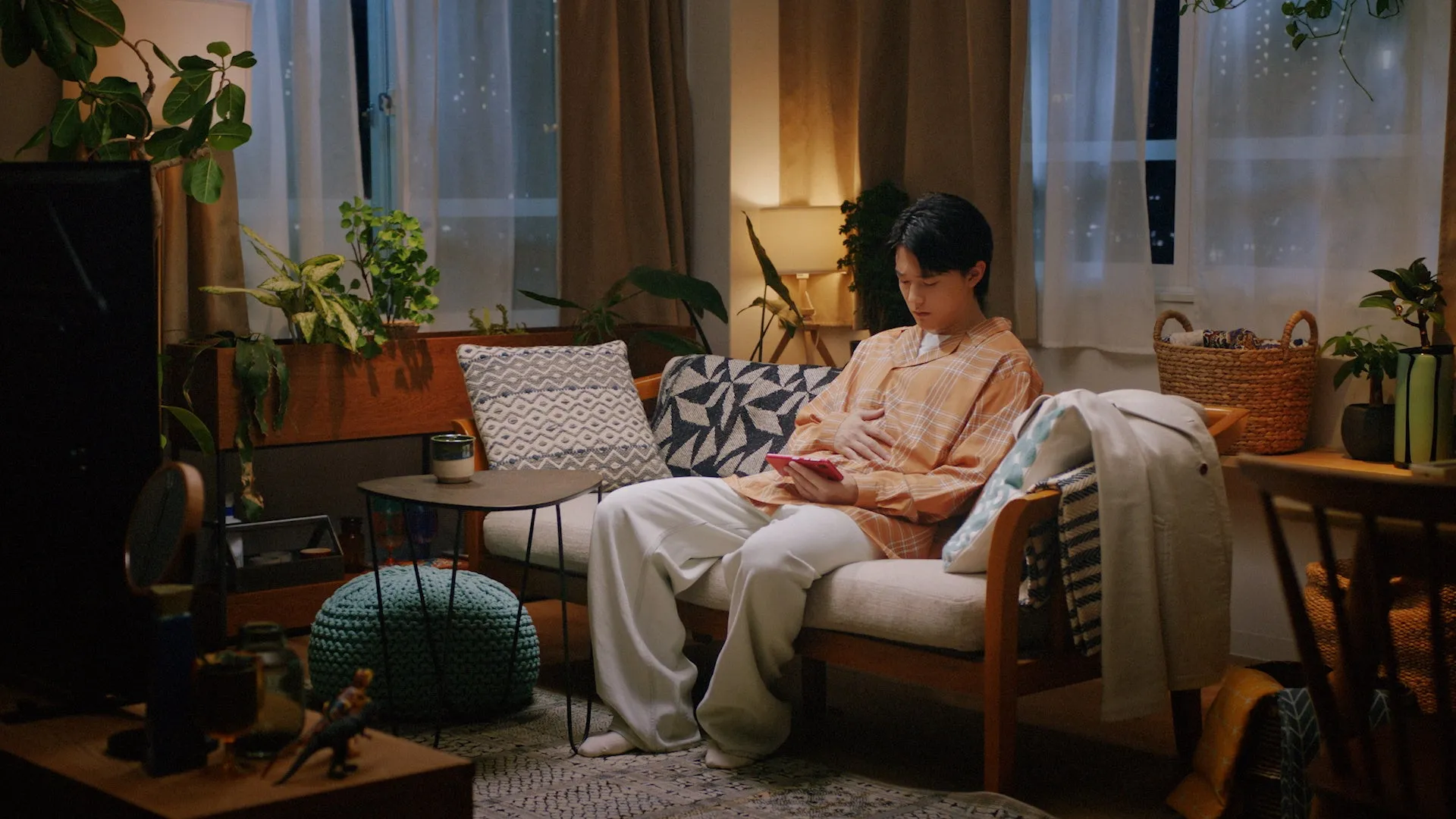 部屋でくつろぐ男性。ソファに座り、スマートフォンを見ている。周りには植物や照明があり、リラックスした雰囲気。