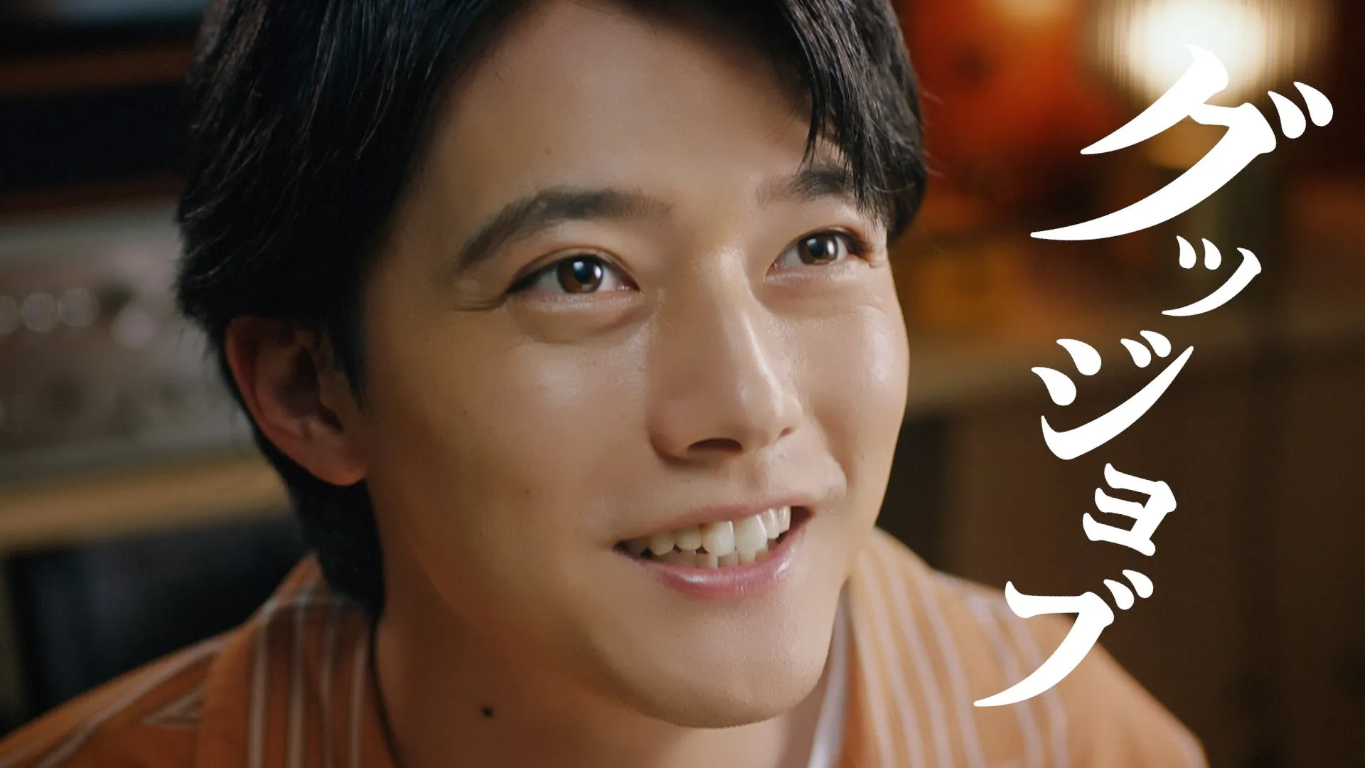 笑顔の男性のクローズアップで、背景にはぼやけた室内シーンがあります。白い文字で「グッジョブ」と書かれています。