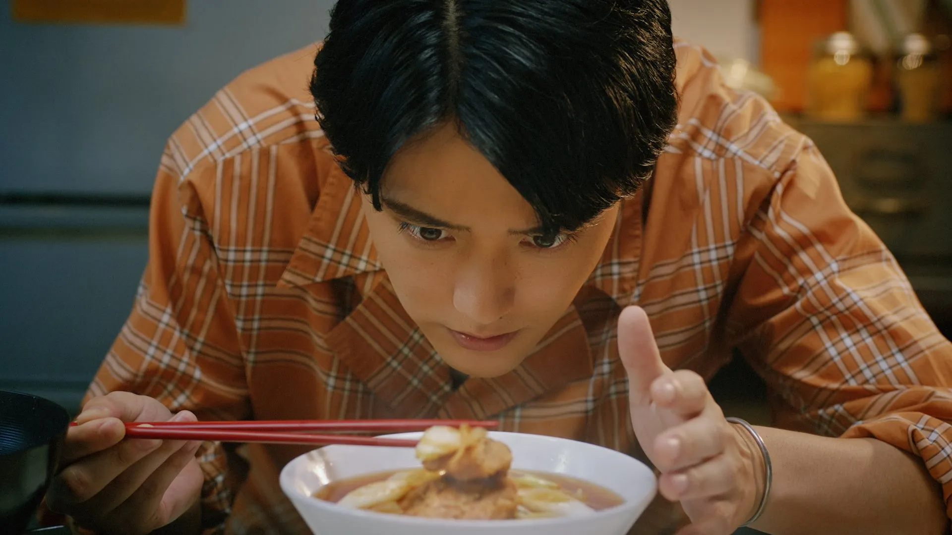 男性がラーメンを食べる様子を描いた写真。彼はオレンジ色のチェック柄のシャツを着ており、箸を使ってラーメンをすくおうとしている。彼の目はラーメンに集中している。