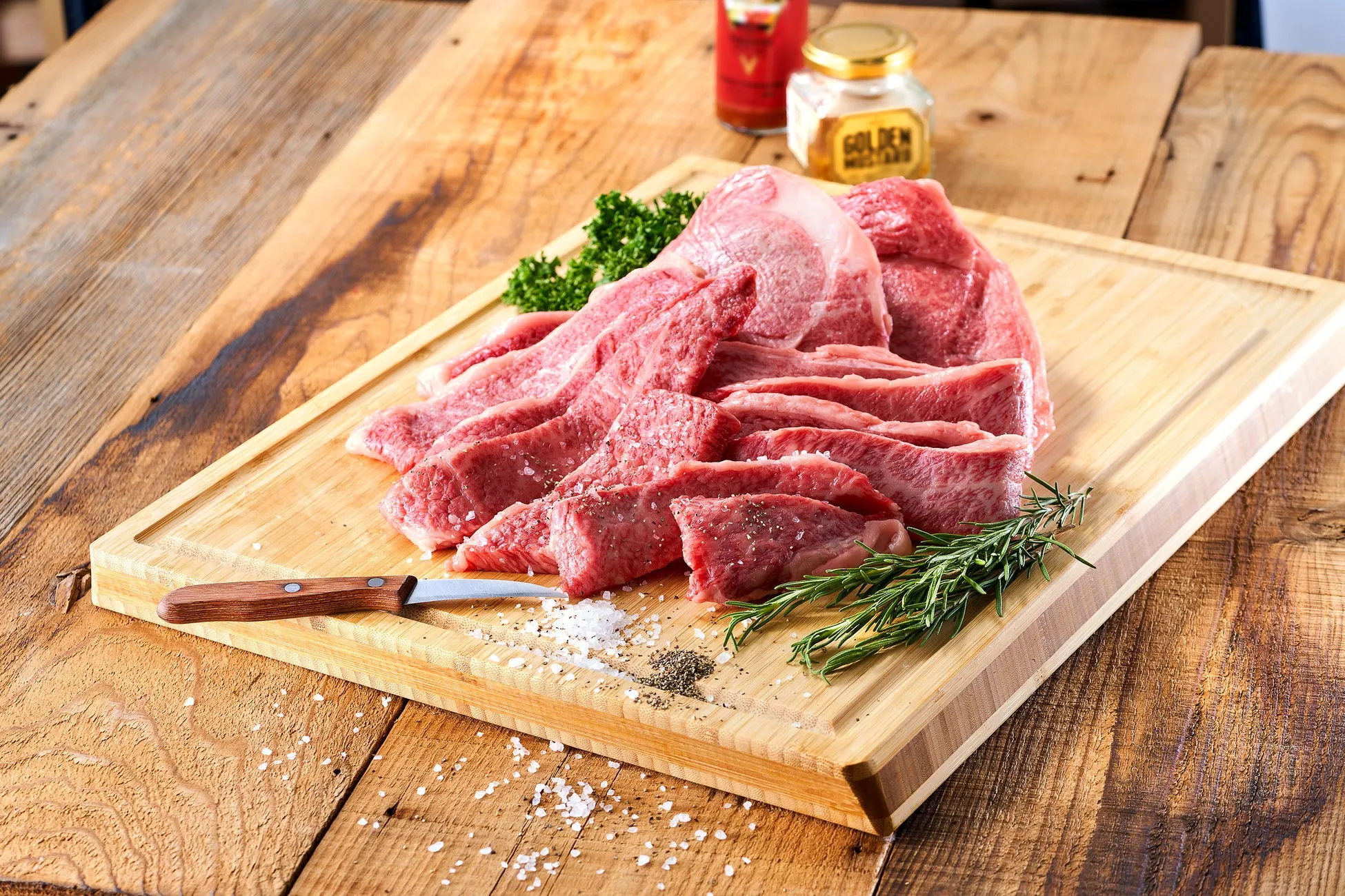 木製のまな板に、カットされた赤身肉とハーブ、調味料が添えられた様子。