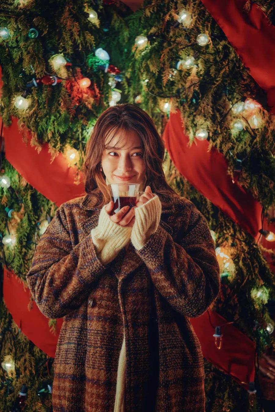 クリスマスツリーを背景に、温かい飲み物を手に微笑む女性。