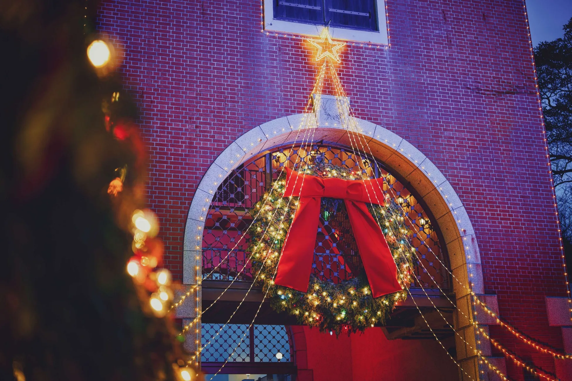 レンガ造りの建物の入り口に飾られたクリスマスリース。大きな赤いリボンと、周りを囲むように飾られた電飾が特徴的。