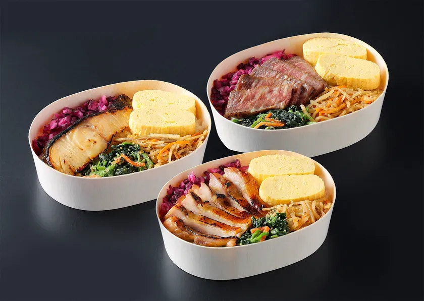 3種類の弁当が並んだ写真。それぞれ白木の容器に入っており、魚、肉、鶏肉がメインで、卵焼き、野菜、ご飯などが詰められている。