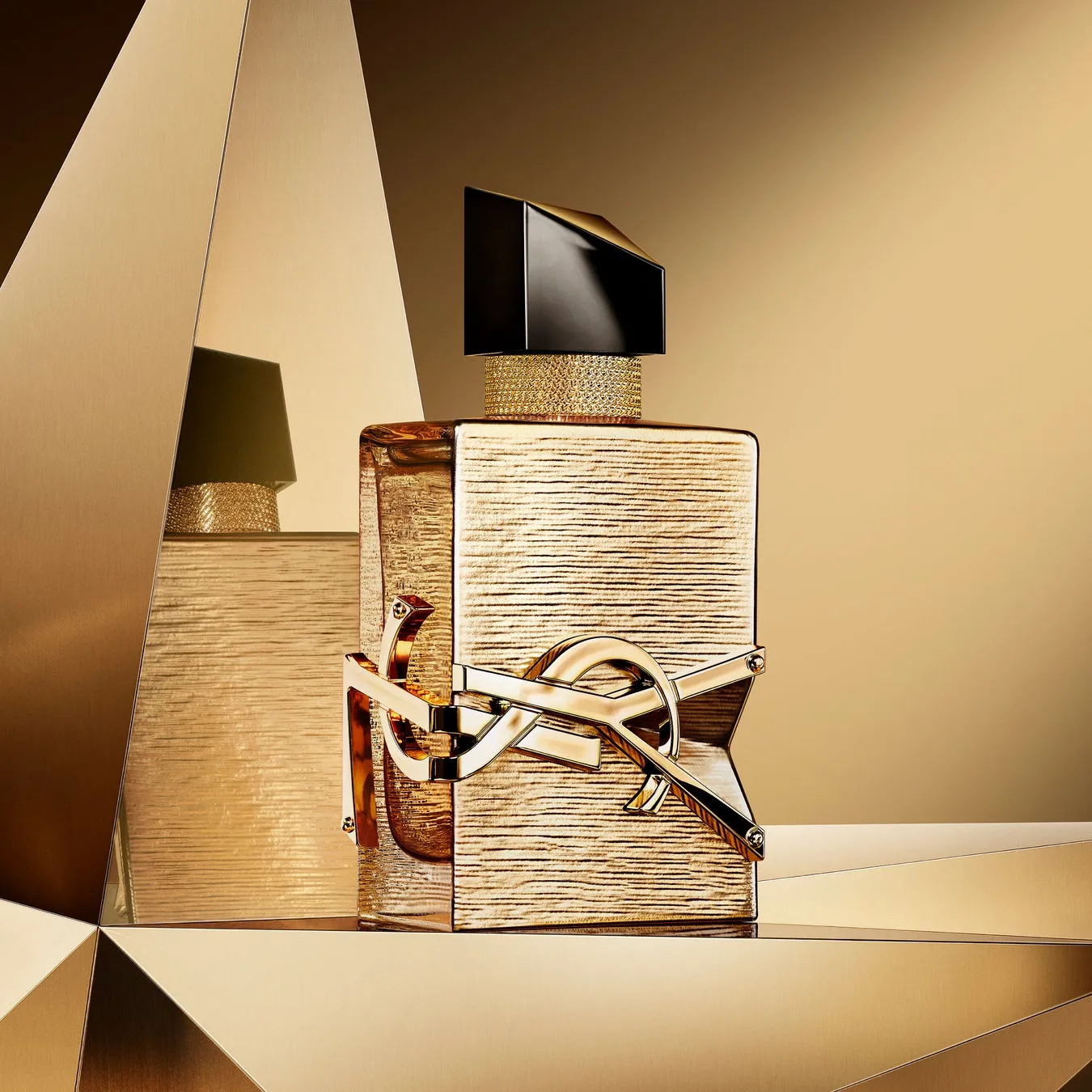 金色の香水ボトルのクローズアップショット。ボトルは四角く、金色の「YSL」のロゴが付いており、背景には黄金の幾何学的な形が配置されています。